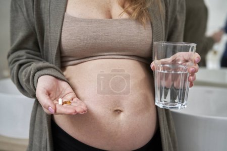 Photo for Caucasian pregnant woman holding nutritional supplements and a glass of water - Royalty Free Image
