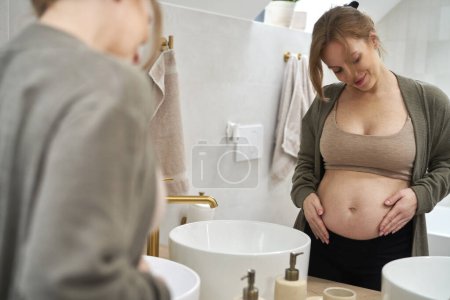 Foto de Caucásica embarazada de pie en el baño y tocando su abdomen - Imagen libre de derechos