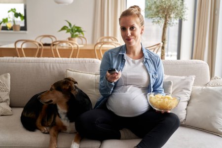 Foto de Mujer embarazada viendo la televisión y comiendo patatas fritas con un perro - Imagen libre de derechos