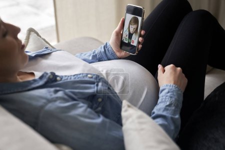 Téléchargez les photos : Femme enceinte allongée sur le canapé et ayant un appel vidéo avec un médecin - en image libre de droit