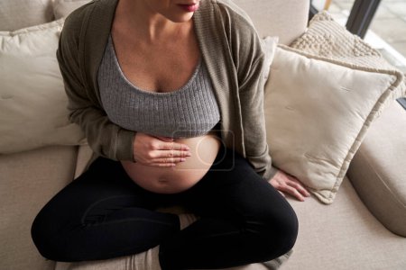 Téléchargez les photos : Femme enceinte caucasienne méconnaissable assise sur un canapé et touchant son abdomen - en image libre de droit