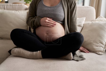 Téléchargez les photos : Vue de face de la femme enceinte assise sur le canapé et touchant son abdomen - en image libre de droit