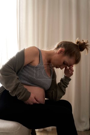 Téléchargez les photos : Femme enceinte anxieuse assise sur le canapé et touchant son abdomen - en image libre de droit