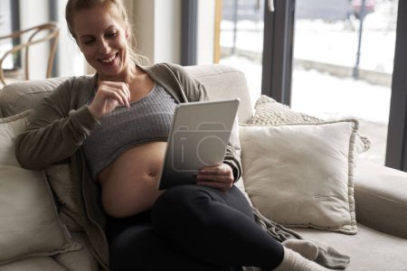 Téléchargez les photos : Femme enceinte caucasienne assise sur un canapé et utilisant une tablette numérique - en image libre de droit
