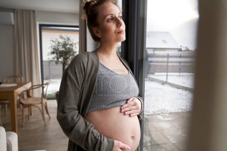 Téléchargez les photos : Femme enceinte anxieuse debout à côté de la fenêtre et touchant son abdomen - en image libre de droit