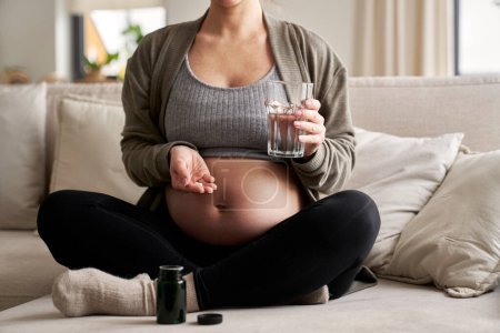 Téléchargez les photos : Femme enceinte caucasienne assise sur le canapé et prenant des suppléments nutritionnels - en image libre de droit