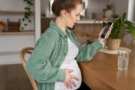 Foto de Mujer embarazada sentada junto a la mesa y recibiendo videollamadas con un médico - Imagen libre de derechos