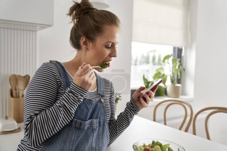 Téléchargez les photos : Femme enceinte manger de la salade et de navigation téléphone mobile - en image libre de droit