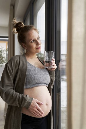 Téléchargez les photos : Femme enceinte anxieuse debout à côté de la fenêtre et touchant son abdomen - en image libre de droit