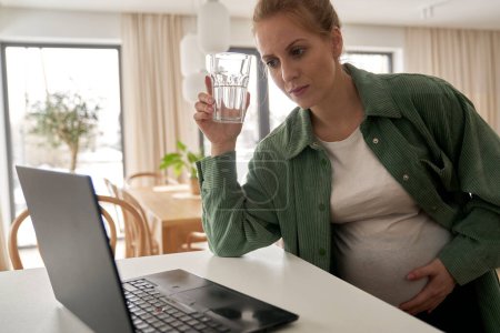 Téléchargez les photos : Femme enceinte utilisant un ordinateur portable dans la cuisine - en image libre de droit