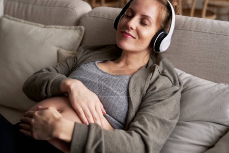 Photo for Caucasian pregnant woman lying down on sofa and listening to music - Royalty Free Image