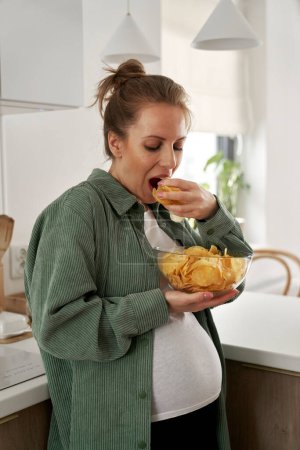 Téléchargez les photos : Femme enceinte chips dans la cuisine - en image libre de droit