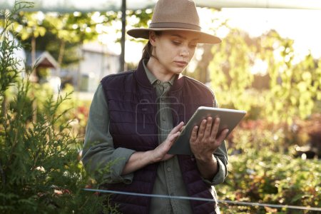 Téléchargez les photos : Femme botaniste caucasienne debout dans la serre et en utilisant une tablette numérique - en image libre de droit