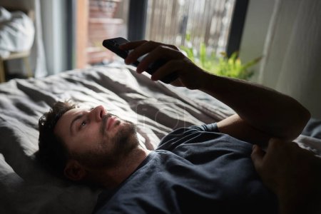 Téléchargez les photos : Homme ennuyé couché sur son lit et naviguant sur son téléphone portable - en image libre de droit