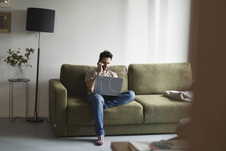Téléchargez les photos : Homme utilisant un ordinateur portable et appelant assis sur le canapé - en image libre de droit