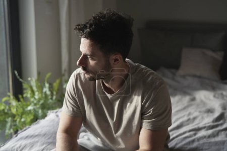 Foto de Hombre ansioso en casa sentado en la cama y mirando hacia otro lado - Imagen libre de derechos