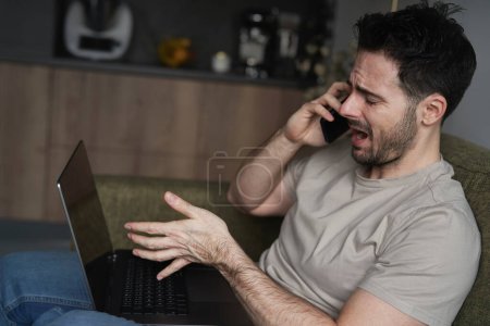 Téléchargez les photos : Homme frustré utilisant un ordinateur portable et appelant assis sur le canapé - en image libre de droit