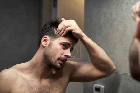 Téléchargez les photos : Homme caucasien vérifier l'état des cheveux dans la salle de bain - en image libre de droit