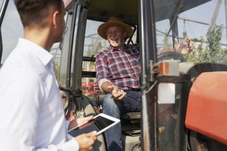 Téléchargez les photos : Fermier senior et représentant des ventes à côté du tracteur parlant sur tablette numérique - en image libre de droit