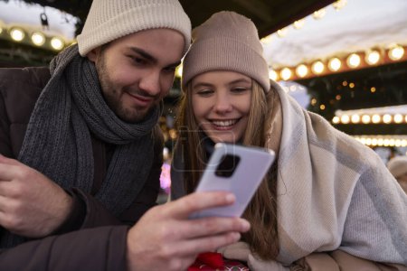Foto de Joven pareja navegando por teléfono juntos en el mercado de Navidad por la noche - Imagen libre de derechos