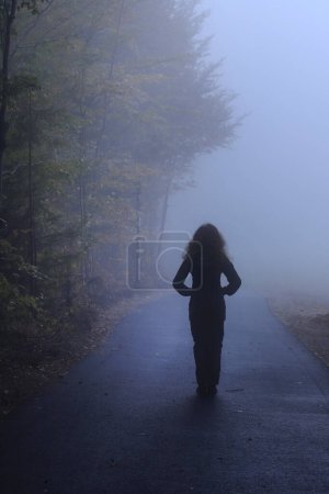 Téléchargez les photos : La femme se tient dans le brouillard, regardant la forêt. Route dangereuse. Ressources graphiques - en image libre de droit