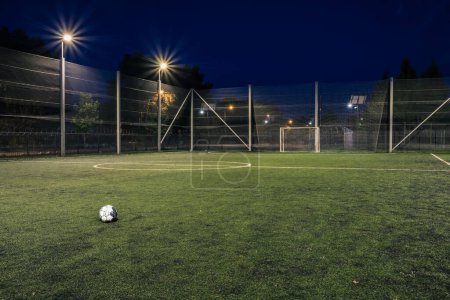 An amateur soccer field with ball illuminated at night. A small football field lit by lanterns in the evening. Green football field illuminated at night. Soccer field in night with spotlight