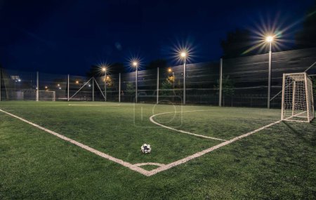 Foto de Un campo de fútbol amateur con pelota iluminada por la noche. Un pequeño campo de fútbol iluminado por linternas en la noche. Campo de fútbol verde iluminado por la noche. Campo de fútbol en la noche con foco - Imagen libre de derechos