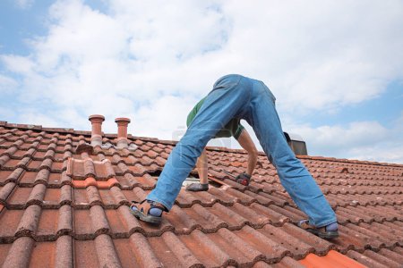 Photo for Worker on the rooftop, replacing broken tiles with new shingles, view from backside - Royalty Free Image