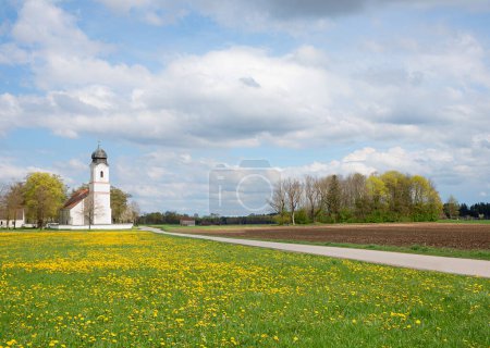 Ländliche Landschaft Hohenkirchen, traditionelle Wallfahrtskirche St. Leonhard, Löwenzahnwiese und Fußweg. Frühling in Oberbayern
