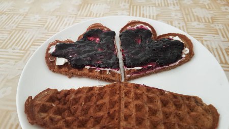 two heart-shaped wholemeal waffles, spread with blueberry jam, on a white porcelain plate. tablecloth with pattern. 