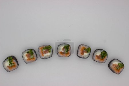 Ten wysokiej rozdzielczości obraz rejestruje ukośny rząd sześciu świeżo wykonanych bułek sushi z łososia, delikatnie umieszczonych na nieskazitelnym białym tle. Kawałki sushi są szczelnie zwinięte z błyszczącą, czarną powierzchnią wodorostów nori, pokrytą perfekcyjnie ugotowaną, whi