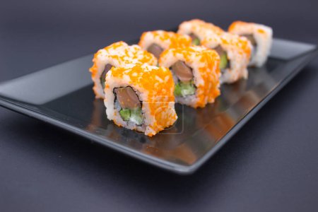 Cette photographie capture une élégante exposition de sushis disposés dans un motif en zigzag ludique sur une plaque d'ardoise sombre. Les morceaux de sushi sont recouverts de masago orange vif, se détachant contre la surface sombre et mate de la plaque. Le contraste est striki
