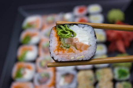 A captivating sushi selection held between chopsticks, poised above an inviting array of various sushi types displayed on a black plate. The scene is framed by a red soy sauce dish and a green wasabi mound, with pickled ginger adding a pop of color. 
