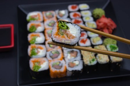 A captivating sushi selection held between chopsticks, poised above an inviting array of various sushi types displayed on a black plate. The scene is framed by a red soy sauce dish and a green wasabi mound, with pickled ginger adding a pop of color. 