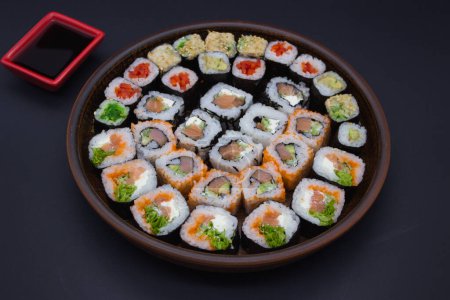 Dieses Bild zeigt eine große runde hölzerne Platte, gefüllt mit einer Reihe von Sushi-Rollen, perfekt in einem kreisförmigen Muster angeordnet. Das Sortiment umfasst eine Auswahl an Brötchen mit frischem Fisch, Avocado und knusprigem Gemüse. Ein kleines rotes Gericht gefüllt wi