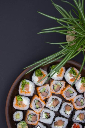This image exudes a sense of Zen with its simple yet stylish presentation of a variety of sushi rolls in a circular wooden tray, positioned next to a potted green plant in a woven basket. The dark background enhances the natural colors of the sushi a
