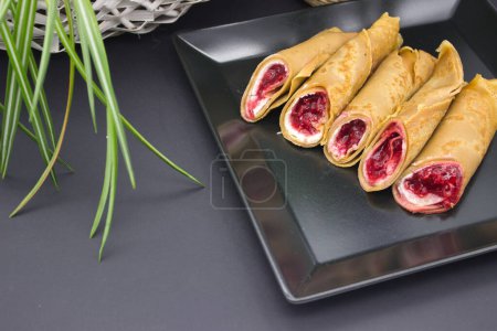 In this appetizing image, a set of golden-brown dessert crepes filled with a luscious berry compote are elegantly arranged on a modern black square plate.