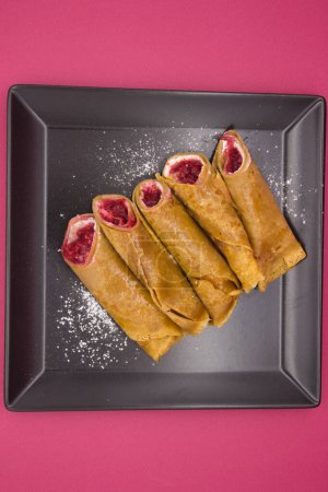 An overhead shot captures the simple elegance of classic crepes filled with a berry mixture, lying on a geometric black plate with a dusting of powdered sugar for garnish. The plate's sharp angles complement the soft curves of the crepes, and the neu