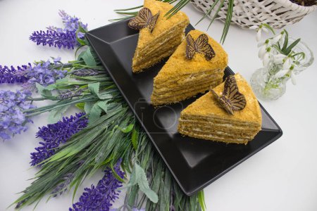 Three honey cake slices adorned with chocolate butterflies, surrounded by fresh flowers.