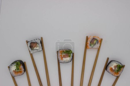 Eine Overhead-Aufnahme, die eine einfallsreiche Sushi-Präsentation zeigt, bei der vier Sushi-Stücke in einer geraden Linie angeordnet sind, jedes flankiert von einem Paar Bambusstäbchen vor einem weißen Hintergrund. Die Sushi-Rollen sind sorgfältig gefertigt, featuri