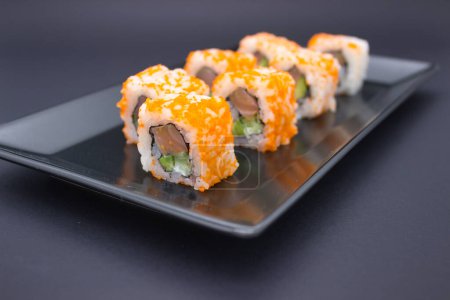 Cette photographie capture une élégante exposition de sushis disposés dans un motif en zigzag ludique sur une plaque d'ardoise sombre. Les morceaux de sushi sont recouverts de masago orange vif, se détachant contre la surface sombre et mate de la plaque. Le contraste est striki