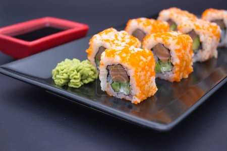 This image features a sleek composition of sushi, wasabi, and soy sauce on a dark, reflective surface. The sushi rolls, topped with bright orange masago, are presented in a line, leading the eye toward a red soy sauce dish in the background. A mound 