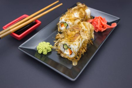 The photograph showcases an exquisite sushi setup, complete with a roll topped with delicate bonito flakes, resting on a black rectangular plate. Accompanying the sushi are vibrant green wasabi and a mound of pickled ginger, with a pair of bamboo cho