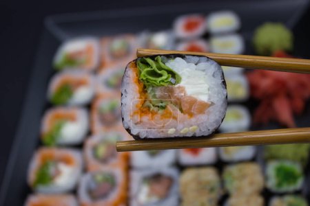 Eine fesselnde Sushi-Auswahl zwischen Essstäbchen, die über einer einladenden Auswahl verschiedener Sushi-Sorten auf einem schwarzen Teller steht. Die Szene wird von einem roten Sojasaucengericht und einem grünen Wasabi-Hügel umrahmt, wobei eingelegter Ingwer einen Farbtupfer setzt.. 