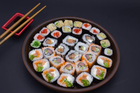 Dieses Bild zeigt eine große runde hölzerne Platte, gefüllt mit einer Reihe von Sushi-Rollen, perfekt in einem kreisförmigen Muster angeordnet. Das Sortiment umfasst eine Auswahl an Brötchen mit frischem Fisch, Avocado und knusprigem Gemüse. Ein kleines rotes Gericht gefüllt wi