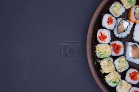 Das Foto zeigt ein kreatives Sushi-Arrangement, das die eine Seite einer runden Holzplatte einnimmt und den dunklen Hintergrund den Rest des Rahmens füllt. Diese exzentrische Komposition schafft einen fesselnden negativen Raum, der das Auge auf das det lenkt
