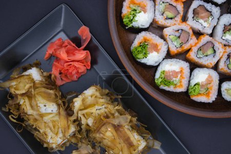 The image artfully captures a sushi feast, featuring two plates with contrasting shapes and textures. On the black rectangular plate, sushi rolls topped with bonito flakes sit alongside pickled ginger, their delicate pink contrasting the golden flake