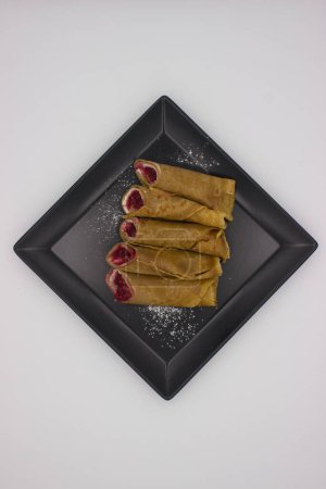 An overhead shot captures the simple elegance of classic crepes filled with a berry mixture, lying on a geometric black plate with a dusting of powdered sugar for garnish. The plate's sharp angles complement the soft curves of the crepes, and the neu