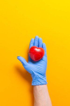 Photo for Hand with blue surgical glove holding a foam heart on orange background. Heart care concept. - Royalty Free Image