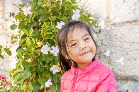 Téléchargez les photos : Petite fille eurasienne chinoise et française souriante très souriante devant un bosquet en fleurs - Portrait - en image libre de droit